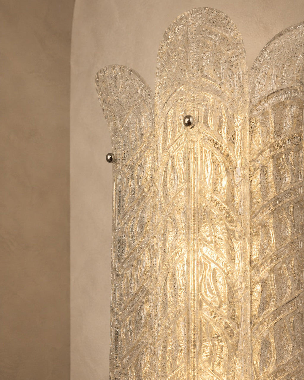 A close-up of a decorative wall sconce or light fixture with an Art Deco style, featuring a textured clear glass or acrylic panel with metallic gold-toned details and small round metal knobs, set against a warm beige plaster wall.