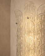 A close-up of a decorative wall sconce or light fixture with an Art Deco style, featuring a textured clear glass or acrylic panel with metallic gold-toned details and small round metal knobs, set against a warm beige plaster wall.