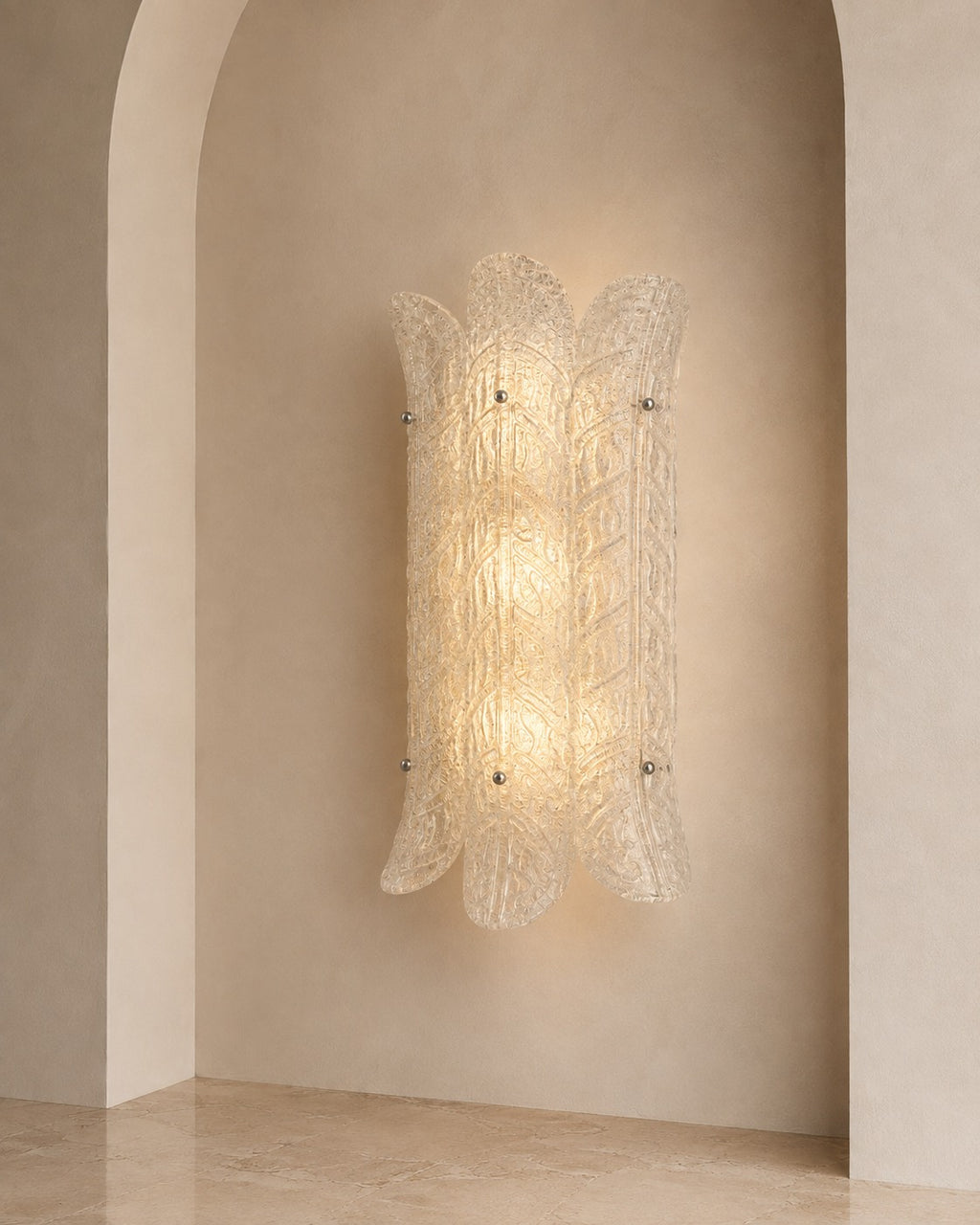 A sculptural wall sconce with a textured, translucent glass shade in a warm glowing light, mounted on a beige plaster wall with an arched niche.