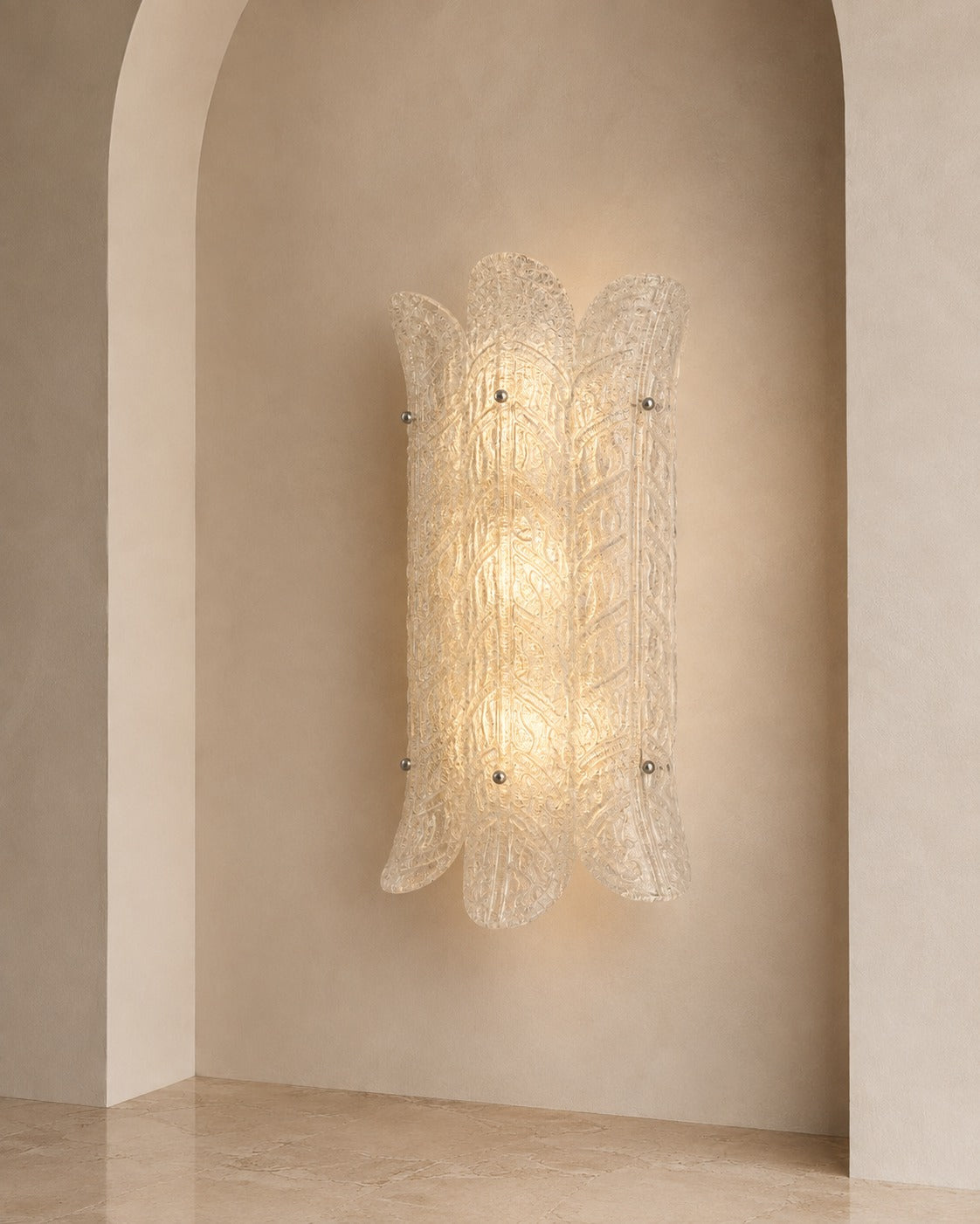 A sculptural wall sconce with a textured, translucent glass shade in a warm glowing light, mounted on a beige plaster wall with an arched niche.