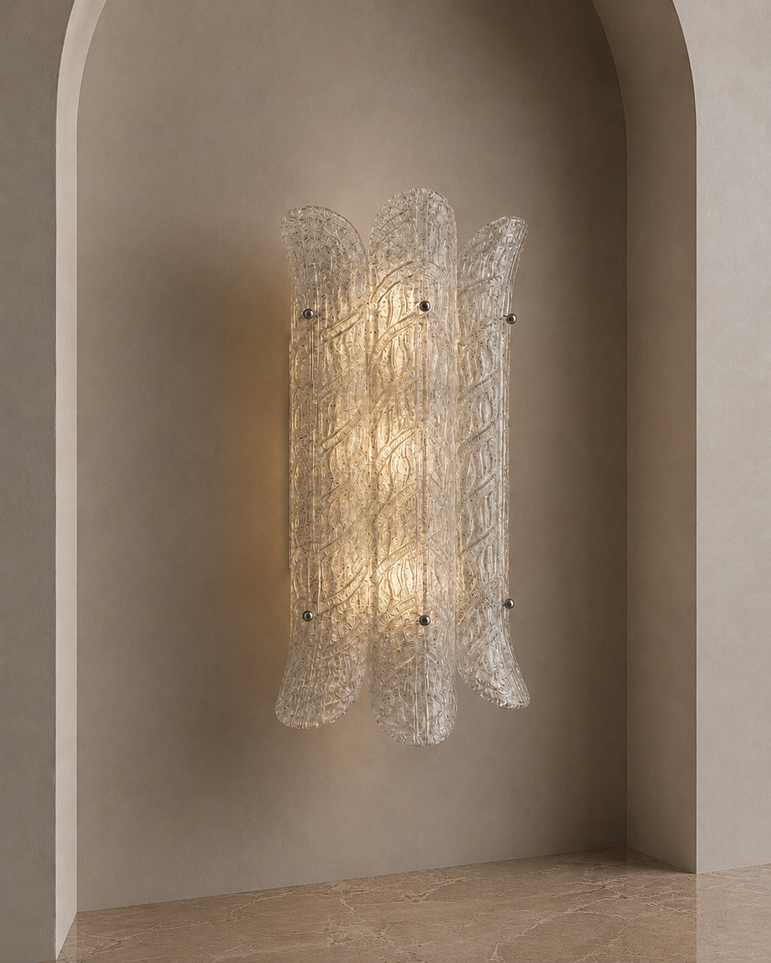 A modern wall sconce with a textured, ribbed clear glass shade, glowing with a warm light, mounted on a beige plaster wall within an arched niche.