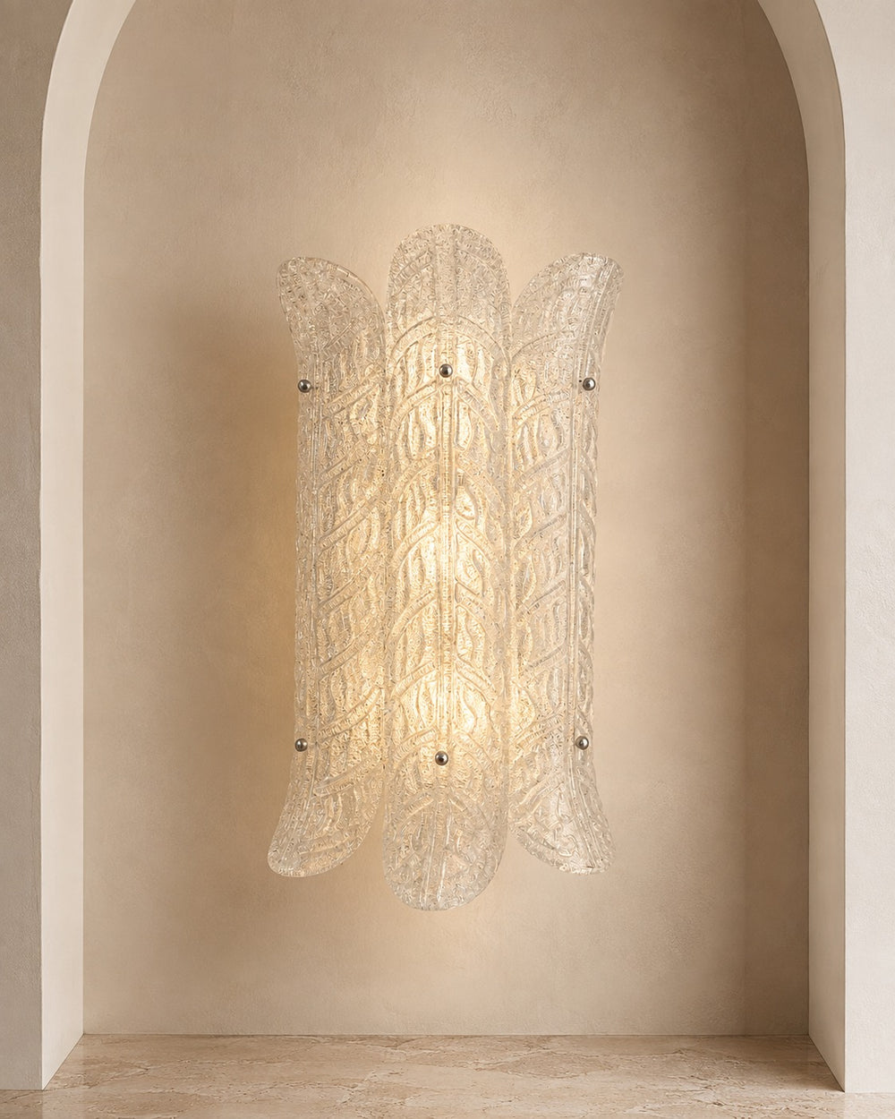 A sculptural wall sconce with a textured, ribbed clear glass shade, mounted on a warm beige plaster wall within an arched niche.