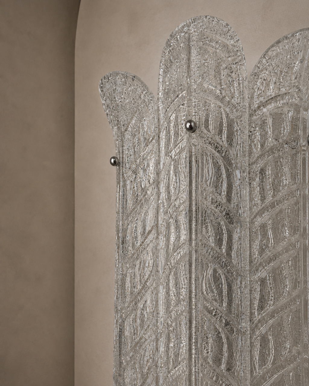 A close-up of a decorative silver-toned screen or divider with an intricate geometric pattern and small metallic knobs, set against a warm beige textured wall.