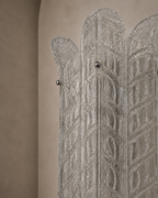 A close-up of a decorative silver-toned screen or divider with an intricate geometric pattern and small metallic knobs, set against a warm beige textured wall.