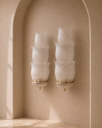 Two matching wall sconces with layered, textured white glass shades and gold-toned metal accents, mounted on a beige plaster wall in an arched alcove.