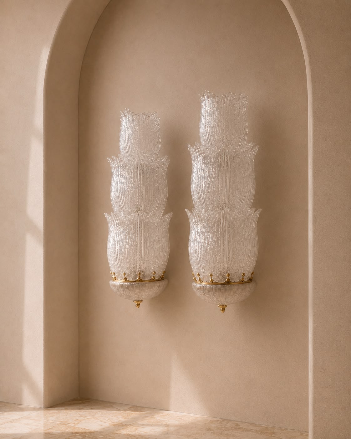 Two matching wall sconces with layered, textured white glass shades and gold-toned metal accents, mounted on a beige plaster wall in an arched alcove.