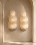 Two matching sculptural wall sconces mounted on a beige plaster wall within an arched alcove. The sconces have layered, petal-like translucent white glass shades with warm yellow light glowing from within, mounted on a textured gray base.