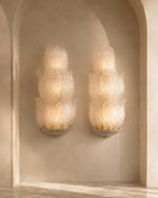 Two matching sculptural wall sconces mounted on a beige plaster wall within an arched alcove. The sconces have layered, petal-like translucent white glass shades with warm yellow light glowing from within, mounted on a textured gray base.