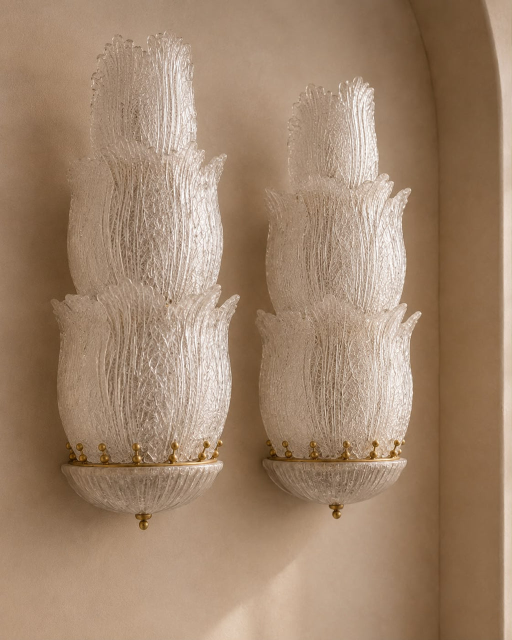 A pair of ornate wall sconces with textured, layered white glass shades and gold metal bases, mounted on a warm beige plaster wall.