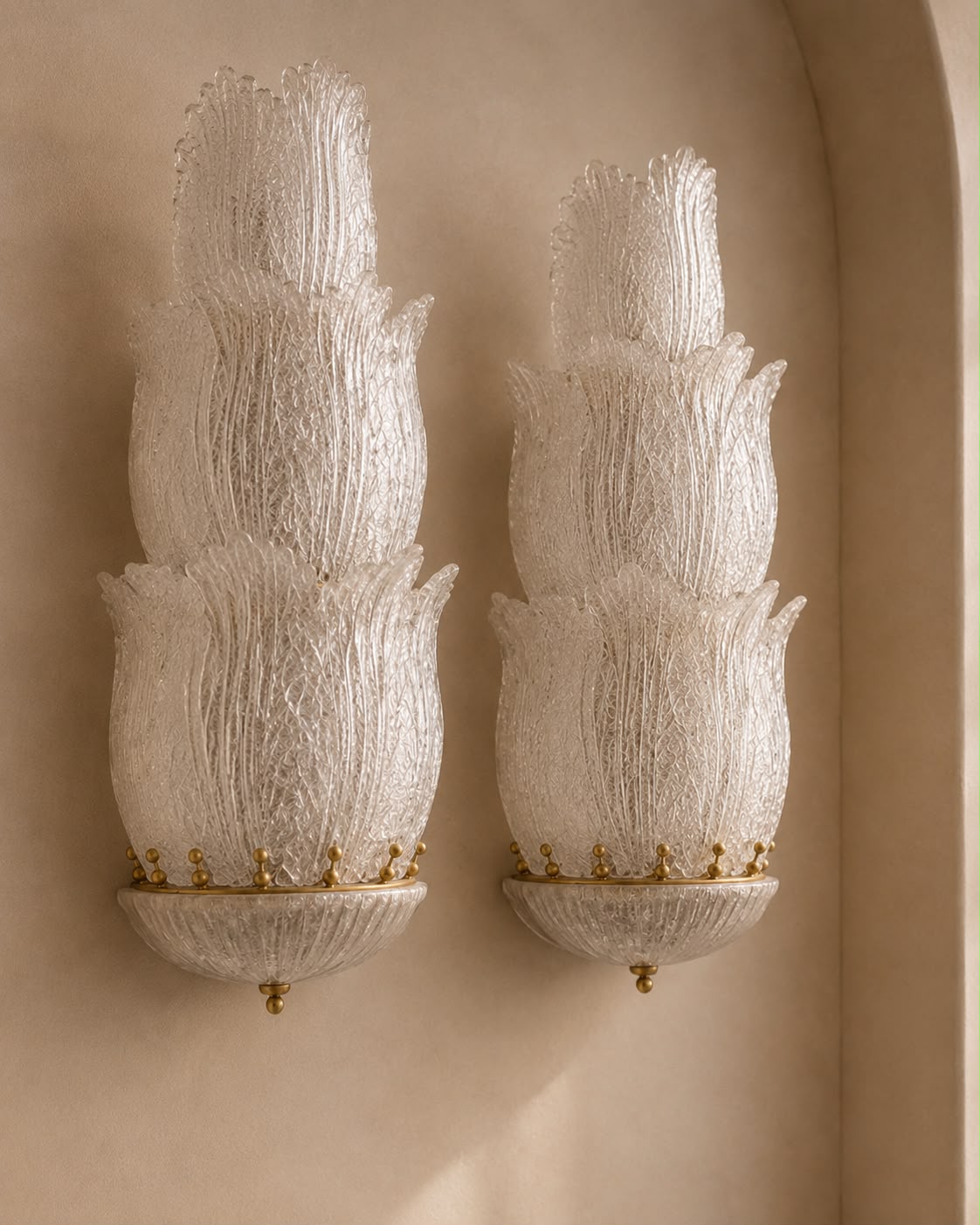 A pair of ornate wall sconces with textured, layered white glass shades and gold metal bases, mounted on a warm beige plaster wall.