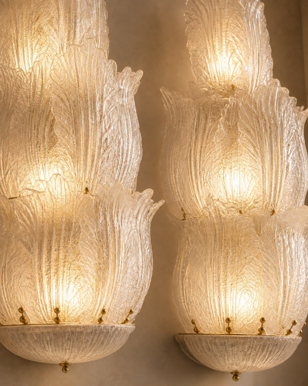 A pair of ornate Murano-style glass wall sconces with layered, petal-like textured glass shades glowing with warm light against a neutral beige wall.