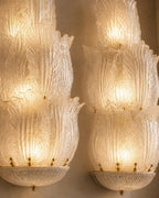 A pair of ornate Murano-style glass wall sconces with layered, petal-like textured glass shades glowing with warm light against a neutral beige wall.