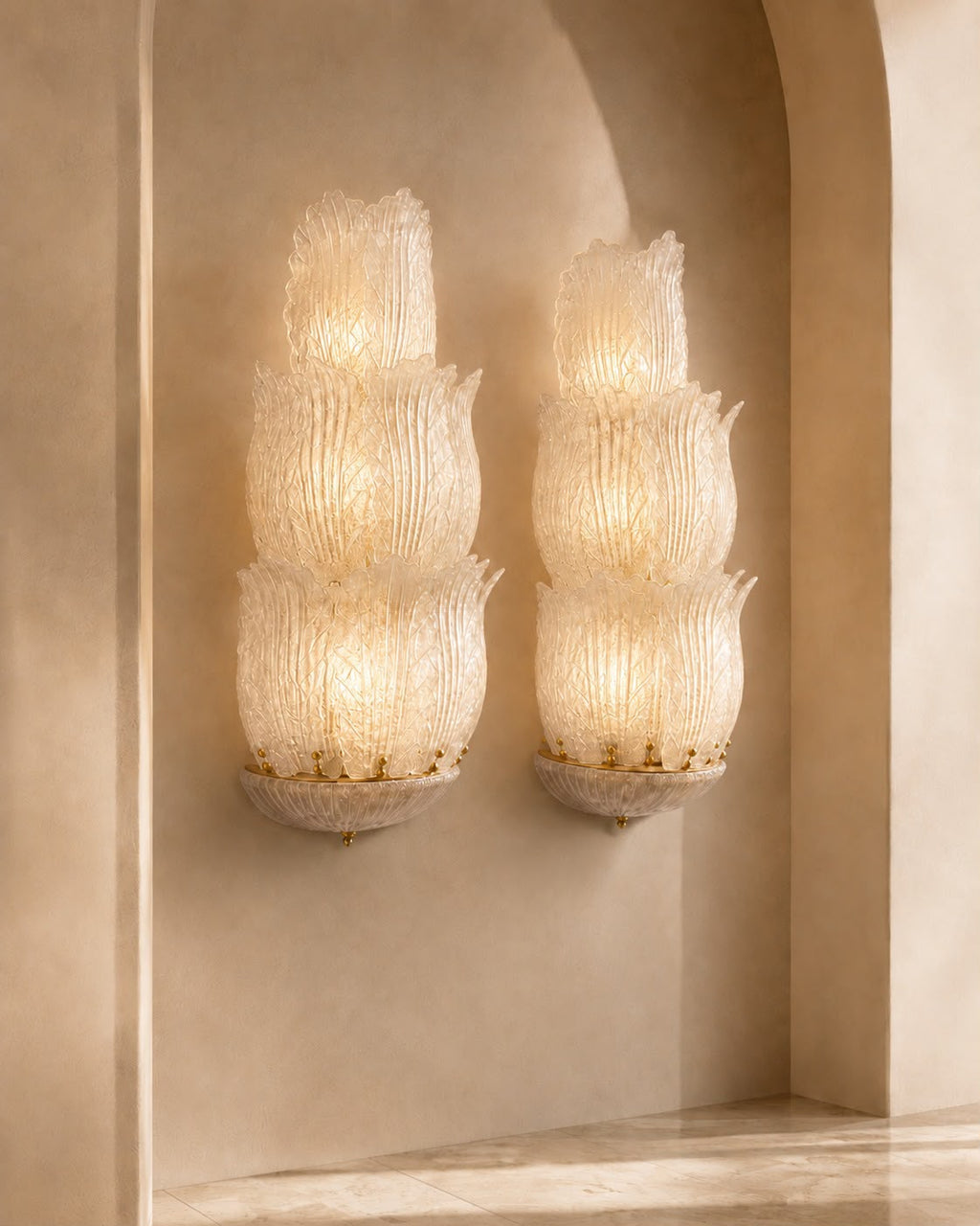 Two matching wall sconces with stacked, textured glass shades glowing warm white, mounted on a beige plaster wall in an arched alcove.