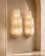 Two matching wall sconces with stacked, textured glass shades glowing warm white, mounted on a beige plaster wall in an arched alcove.