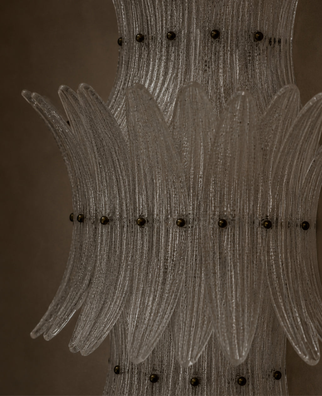 A close-up of a clear ribbed glass chandelier or decorative light fixture with multiple curved glass arms and small dark metal accents against a warm brown background.
