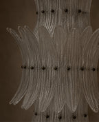 A close-up of a clear ribbed glass chandelier or decorative light fixture with multiple curved glass arms and small dark metal accents against a warm brown background.