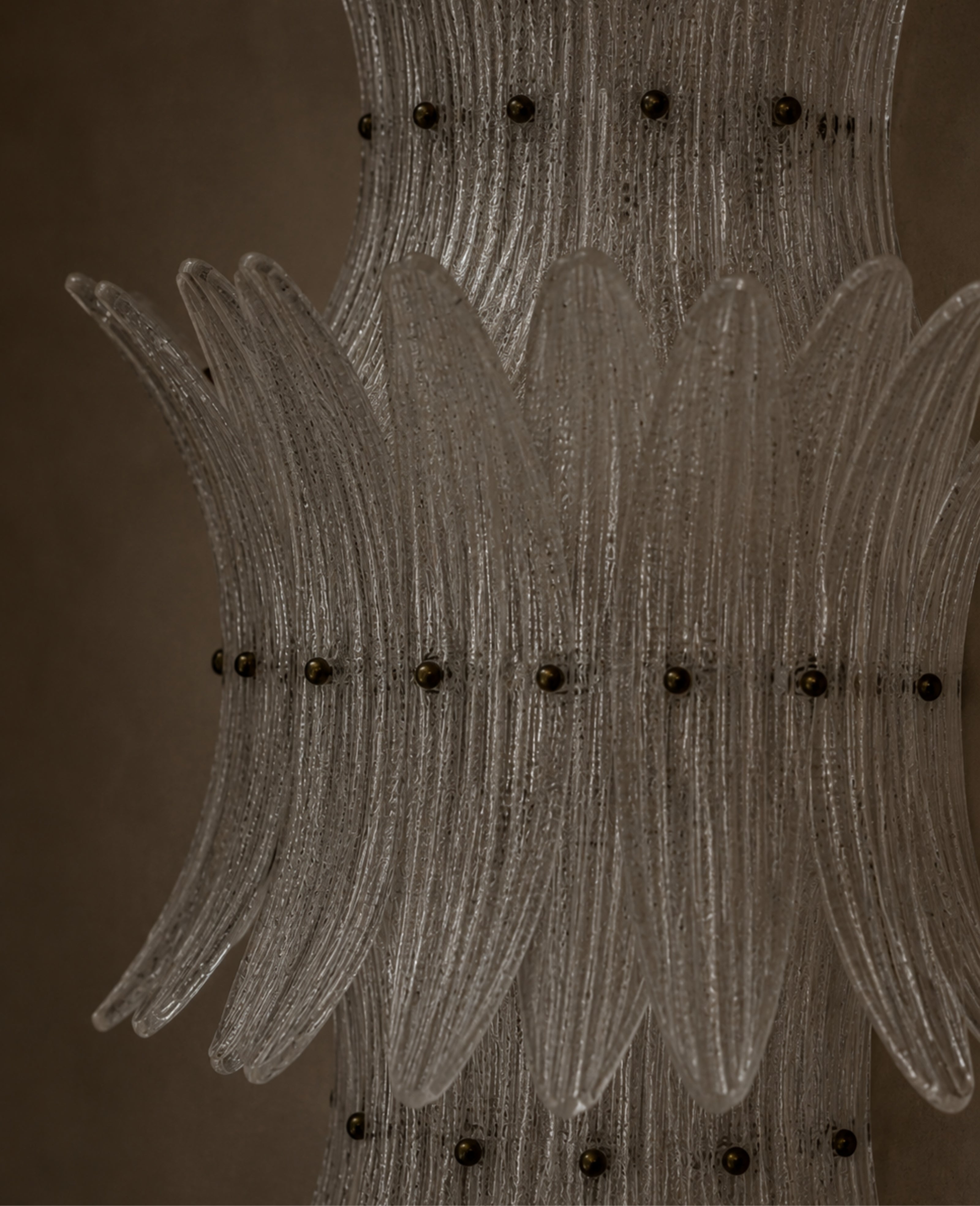 A close-up of a clear ribbed glass chandelier or decorative light fixture with multiple curved glass arms and small dark metal accents against a warm brown background.