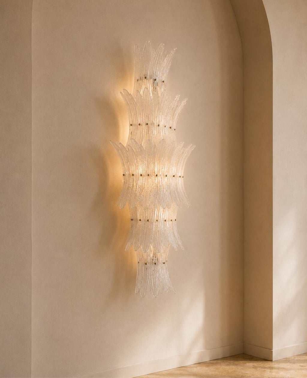 A modern wall-mounted chandelier with layered, translucent glass or acrylic petal-like elements and warm integrated lighting, shown mounted on a beige plaster wall in an arched interior corner.