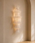 A modern wall-mounted chandelier with layered, translucent glass or acrylic petal-like elements and warm integrated lighting, shown mounted on a beige plaster wall in an arched interior corner.