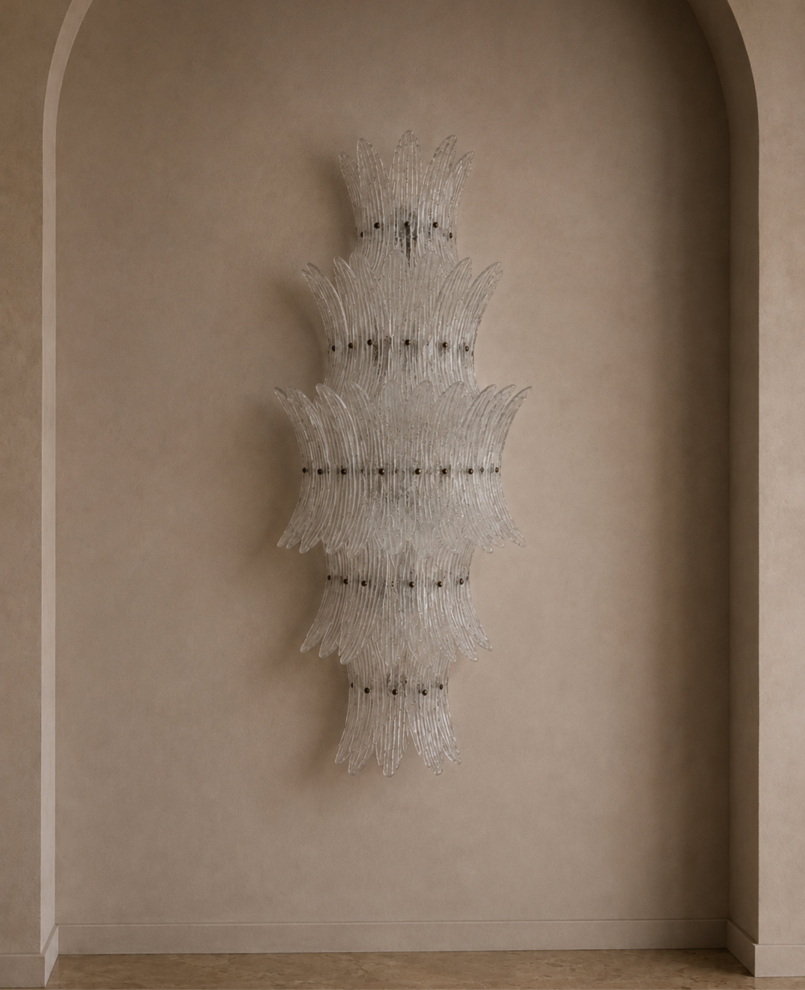 A large, sculptural wall-mounted chandelier made of clear ribbed glass tubes arranged in layered, fan-like shapes, hanging against a warm beige plaster wall with an arched architectural frame.