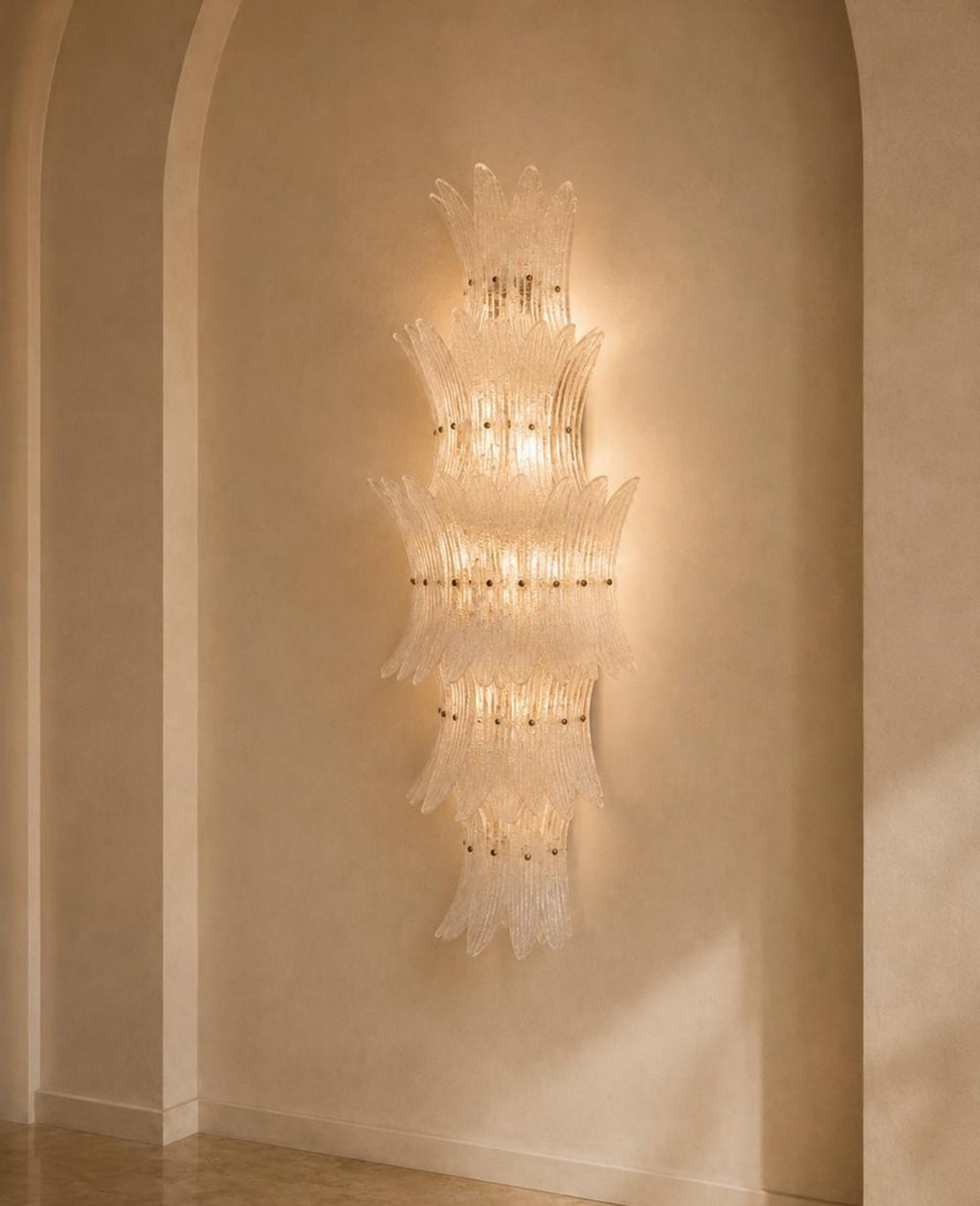 A tall, multi-tiered Murano-style glass chandelier wall sconce with a warm glowing light, mounted on a beige plaster wall in an arched alcove.