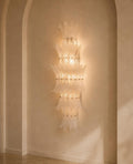 A tall, multi-tiered Murano-style glass chandelier wall sconce with a warm glowing light, mounted on a beige plaster wall in an arched alcove.