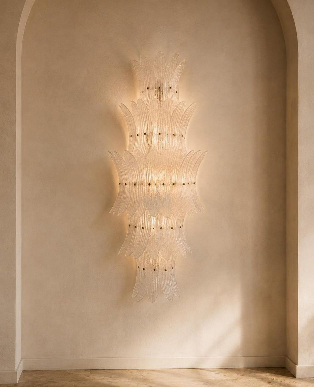 A sculptural, tiered chandelier made of clear glass rods, illuminated with a warm golden glow, mounted on a textured beige plaster wall in an arched alcove.