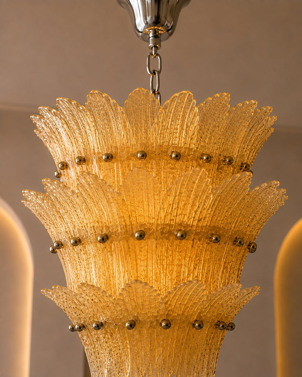 A close-up of a tiered glass chandelier shade made of textured yellow-gold glass with small metallic gold finials, hanging from a silver chain.