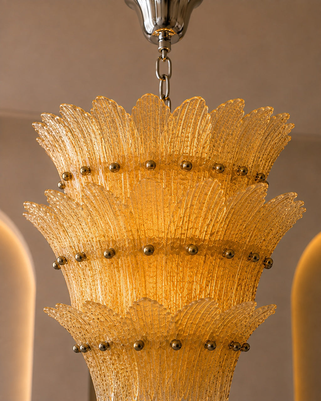 A close-up of a tiered glass chandelier shade made of textured yellow-gold glass with small metallic gold finials, hanging from a silver chain.