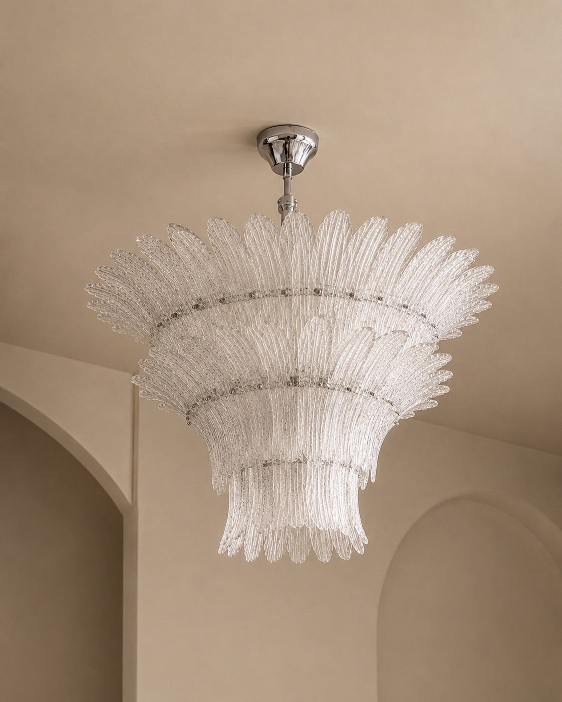 A modern chandelier with a silver ceiling canopy and a tiered, sunburst-style clear glass shade, shown hanging in a beige interior room.