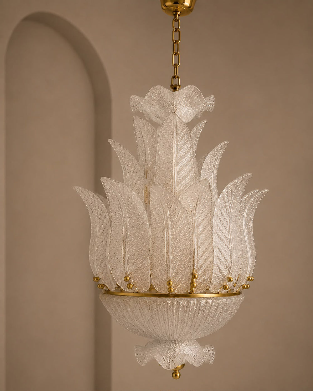 A decorative hanging chandelier with layered translucent white petal or leaf-shaped glass shades and gold-toned metal hardware, suspended against a warm beige wall with an arched architectural detail.