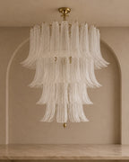 A tiered chandelier with cascading textured glass shades and gold-toned hardware, suspended in front of a beige arched wall.