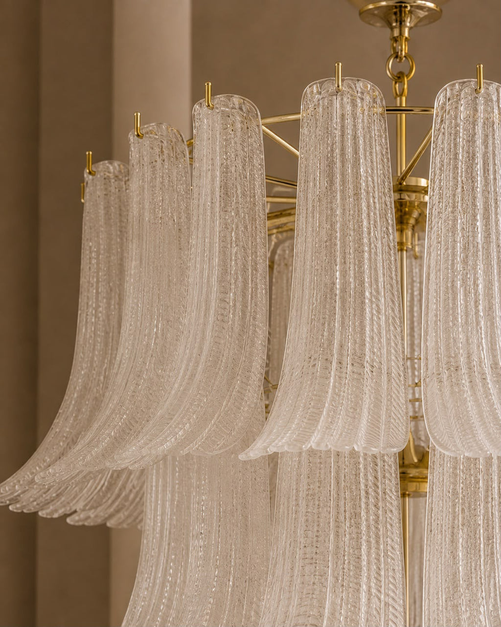 A close-up of a modern chandelier featuring multiple curved, translucent textured glass shades suspended from a polished gold brass frame.