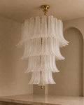 A large tiered chandelier with cascading translucent white glass shades and gold-toned metal hardware, hanging in a warm beige interior room.