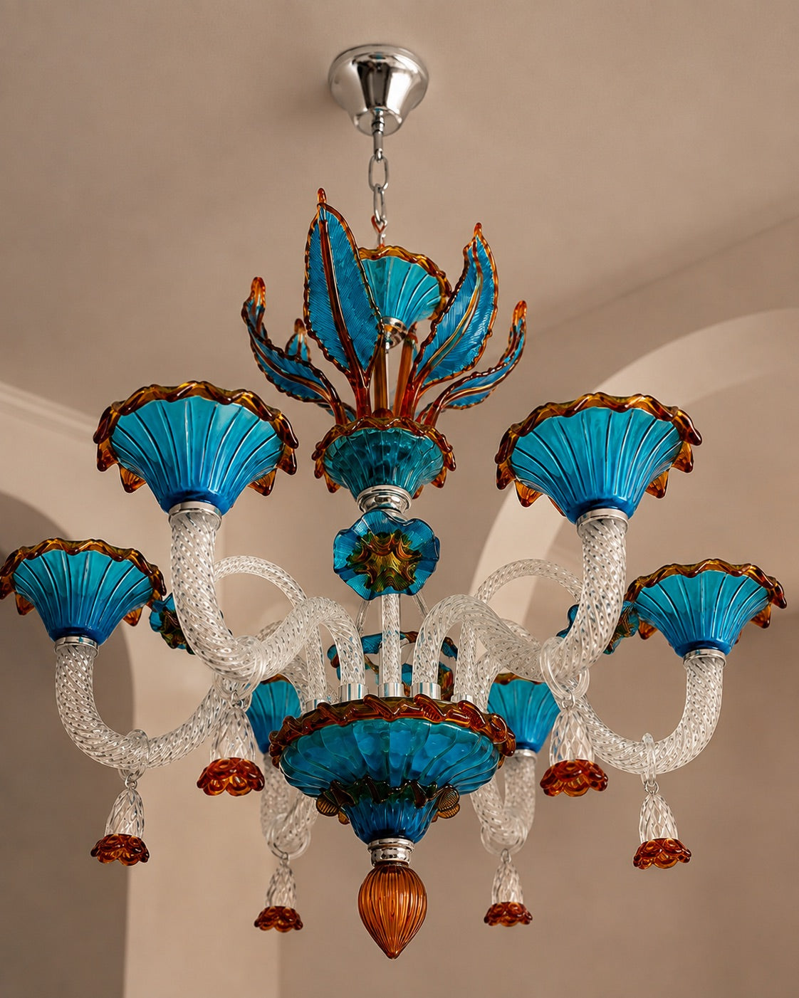 A decorative chandelier with six turquoise blue glass flower-like shades, clear textured glass arms, and amber-brown accents, hanging from a silver ceiling canopy in an indoor room.