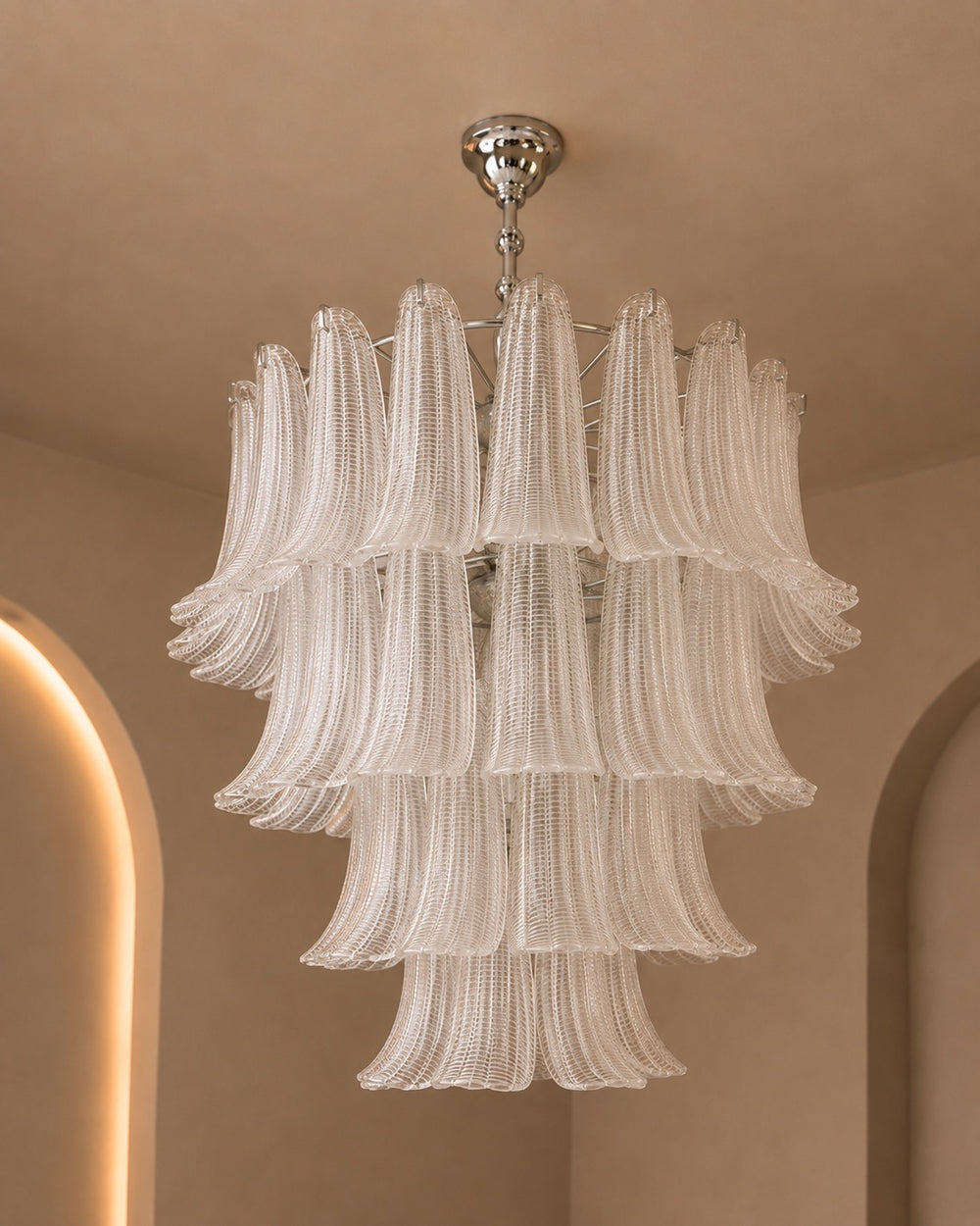 A modern multi-tiered chandelier with textured, fluted glass shades in a warm off-white or cream tone, suspended from a polished silver ceiling mount against a beige plaster wall with arched architectural details.