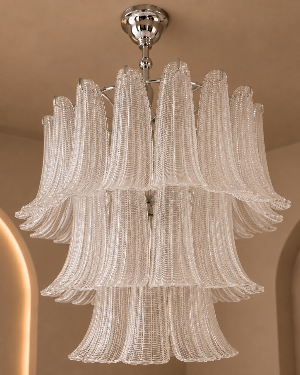 A multi-tiered chandelier with textured, fluted glass shades in a warm off-white or cream tone, suspended from a polished silver-toned metal frame and ceiling mount against a beige wall background.