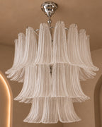A multi-tiered chandelier with textured, fluted glass shades in a warm off-white or cream tone, suspended from a polished silver-toned metal frame and ceiling mount against a beige wall background.