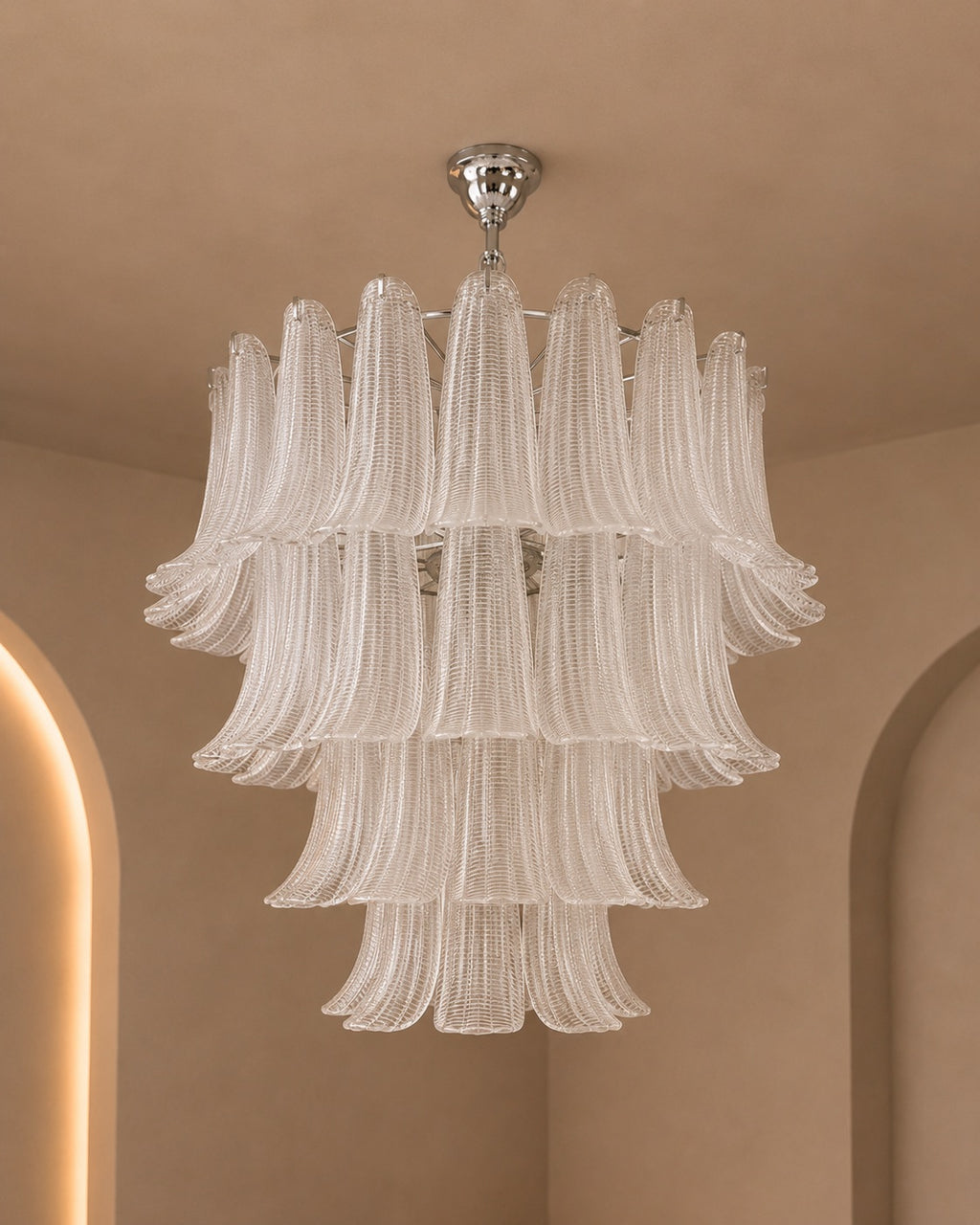 A modern tiered chandelier with fluted clear glass shades and a polished silver ceiling mount, shown hanging in a warm beige interior with arched architectural details.