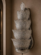 A tiered crystal wall sconce with five stacked textured glass bowls on a polished silver metal backing, mounted on a warm brown wall.