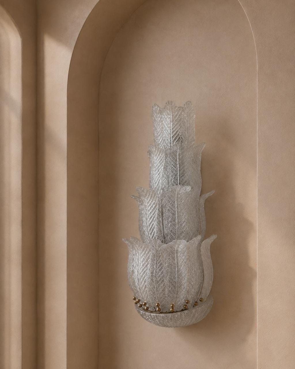A sculptural, tiered wall sconce made of textured, translucent white glass, mounted on a warm beige plaster wall with an arched niche.