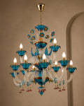 An ornate multi-light chandelier with turquoise blue glass floral elements, gold-toned metal hardware, and clear glass crystal arms, shown hanging against a beige wall.