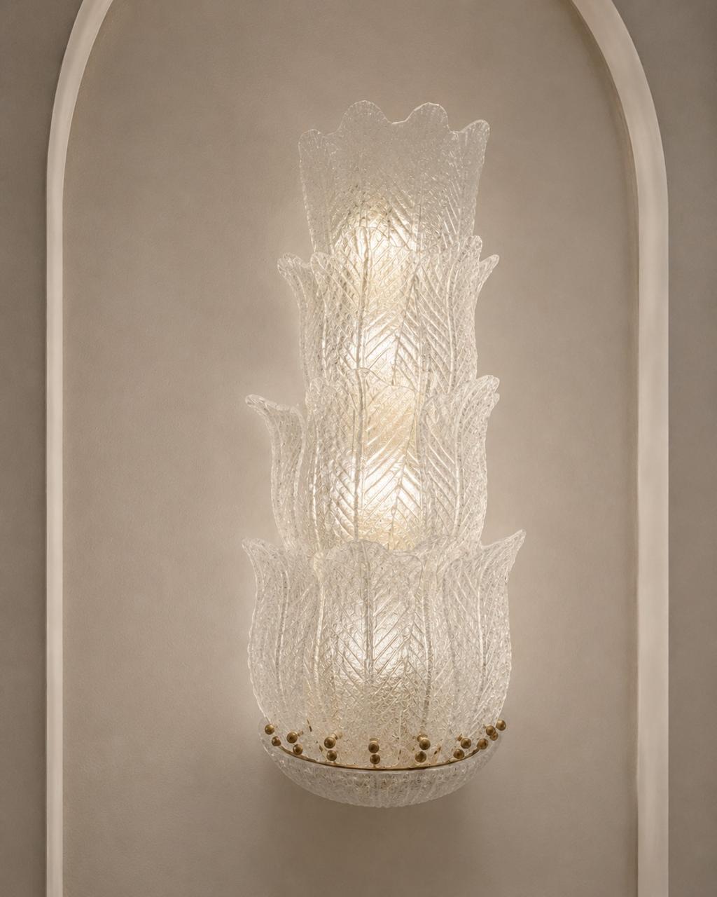 A sculptural, multi-tiered wall sconce made of textured, translucent white glass, mounted on a warm beige wall within a white arched niche.
