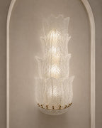 A sculptural, multi-tiered wall sconce made of textured, translucent white glass, mounted on a warm beige wall within a white arched niche.