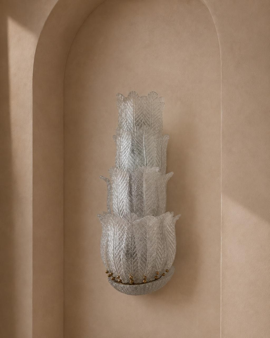 A sculptural, multi-tiered wall sconce made of textured, translucent frosted glass, mounted on a warm beige plaster wall in an arched niche.