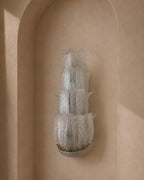 A sculptural, multi-tiered wall sconce made of textured, translucent frosted glass, mounted on a warm beige plaster wall in an arched niche.