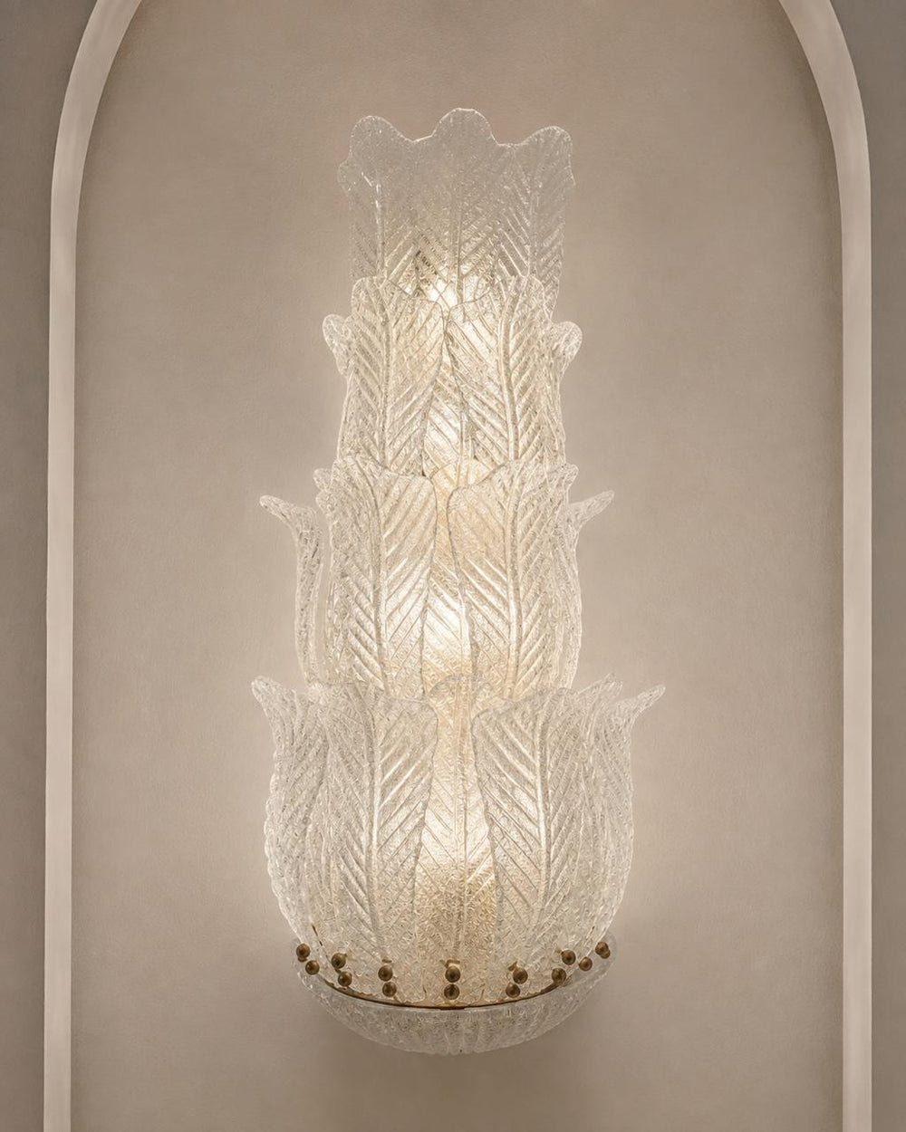 A tiered Murano-style glass wall sconce with textured, leaf-like ribbed glass and a warm glowing light, mounted on a beige wall with an arched architectural frame.