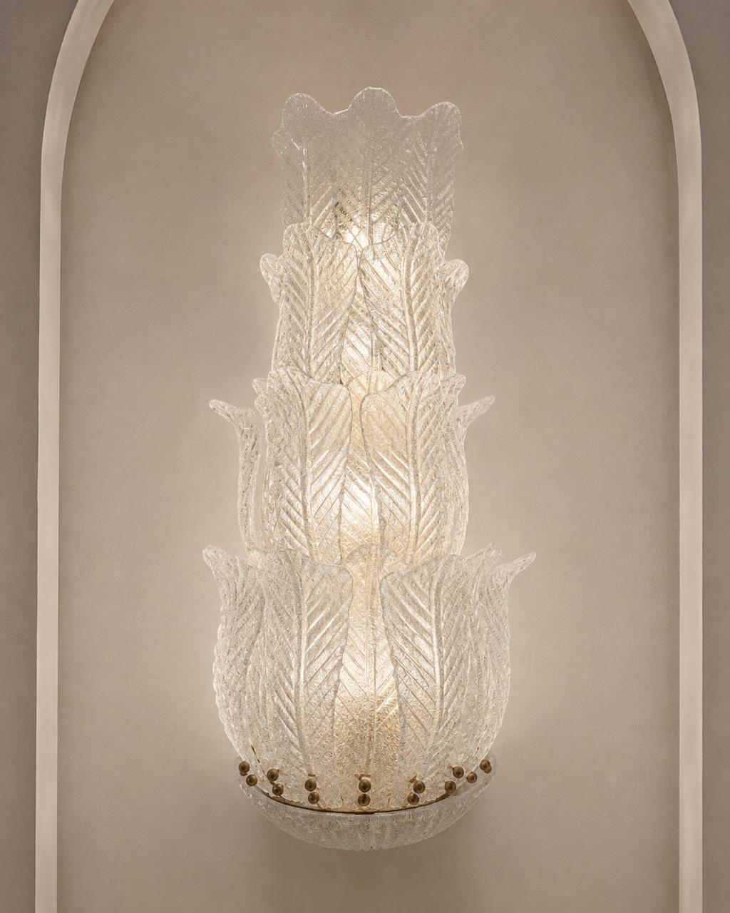 A tiered Murano-style glass wall sconce with textured, leaf-like ribbed glass and a warm glowing light, mounted on a beige wall with an arched architectural frame.