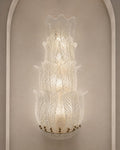A tiered Murano-style glass wall sconce with textured, leaf-like ribbed glass and a warm glowing light, mounted on a beige wall with an arched architectural frame.