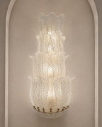 A tiered Murano-style glass wall sconce with textured, leaf-like ribbed glass and a warm glowing light, mounted on a beige wall with an arched architectural frame.
